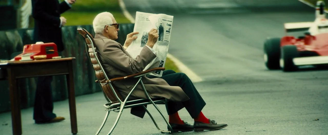 Rush (2013) movie still: fullbody — An elderly man in a tan coat sits on a racetrack tarmac. He is reading a newspaper while a race car sits in the…