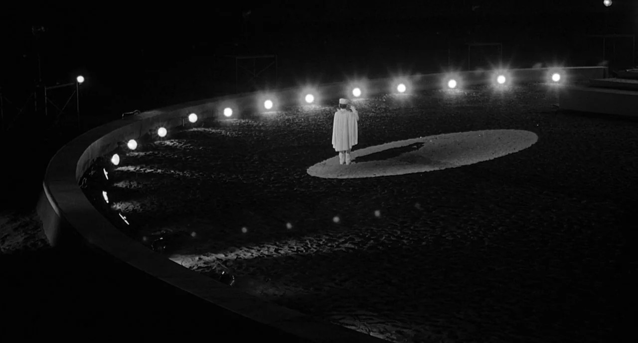 8 1/2 (1963) movie still: wide — A dark, dirt-covered circular performance ring is illuminated by a curved line of lights. A single person dressed…