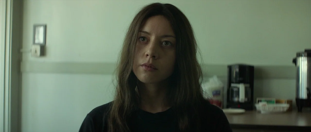 Ingrid Goes West (2017) movie still: medium — A spare breakroom features plain walls, a coffee maker, and an urn on a table. A woman with long brown…