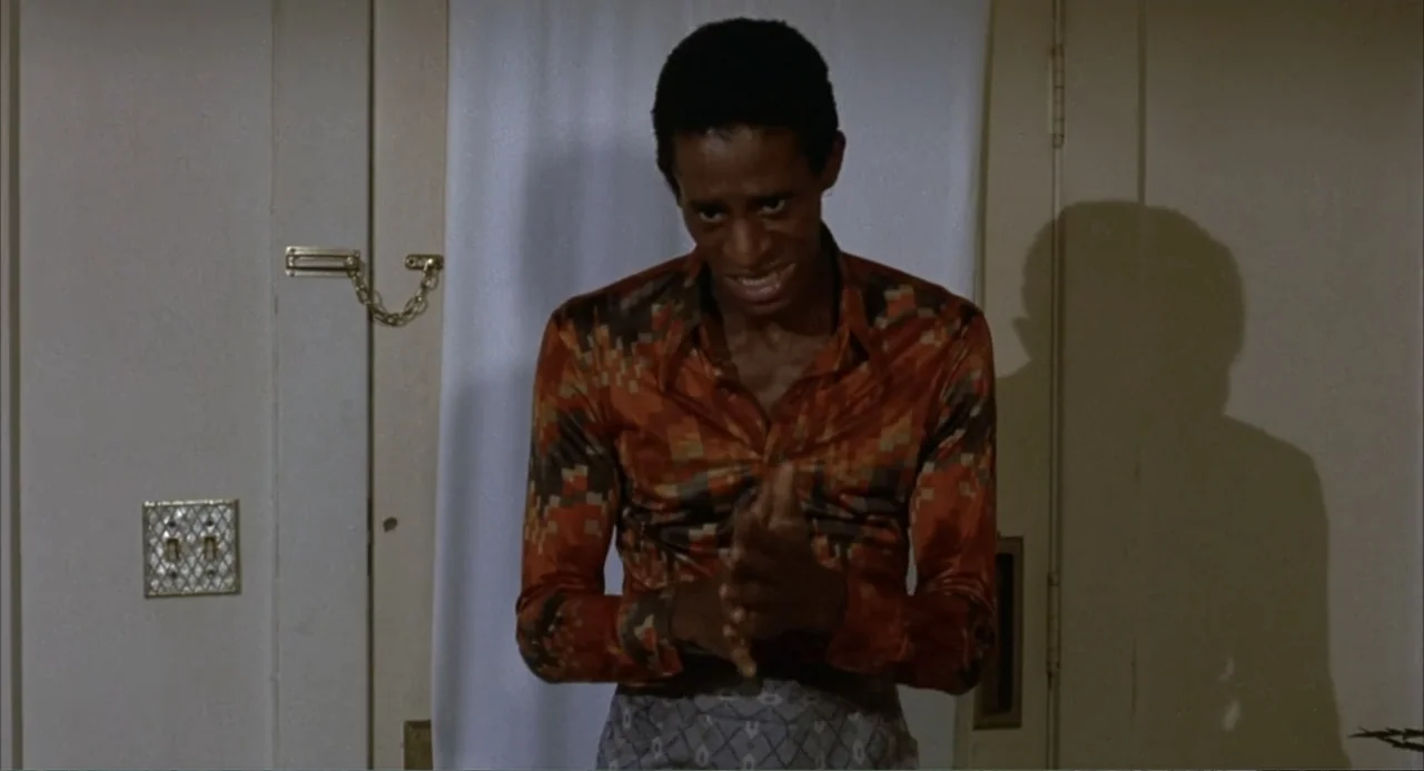 Foxy Brown (1974) movie still: medium — A man stands indoors against a plain door featuring a gold security chain lock. The subject clasps his hands…