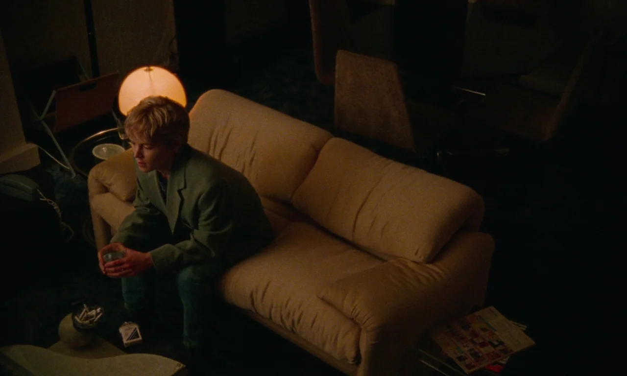 Blue Jean (2022) movie still: medium — A dark living room features a beige sofa and a small round table. A young man sits alone on the edge of the…