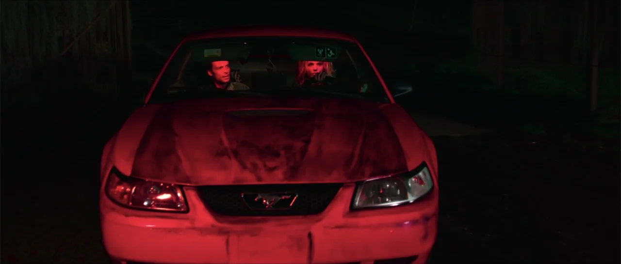 68 Kill (2017) movie still: medium — A dark night setting features the front view of a red Ford Mustang parked. Two people are visible through the…