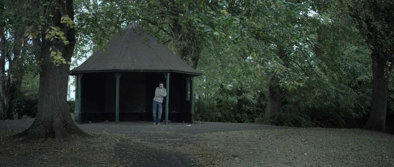 What Richard Did (2012) movie still: wide — A secluded park features a small dark-painted wooden pavilion surrounded by mature trees. A lone young…