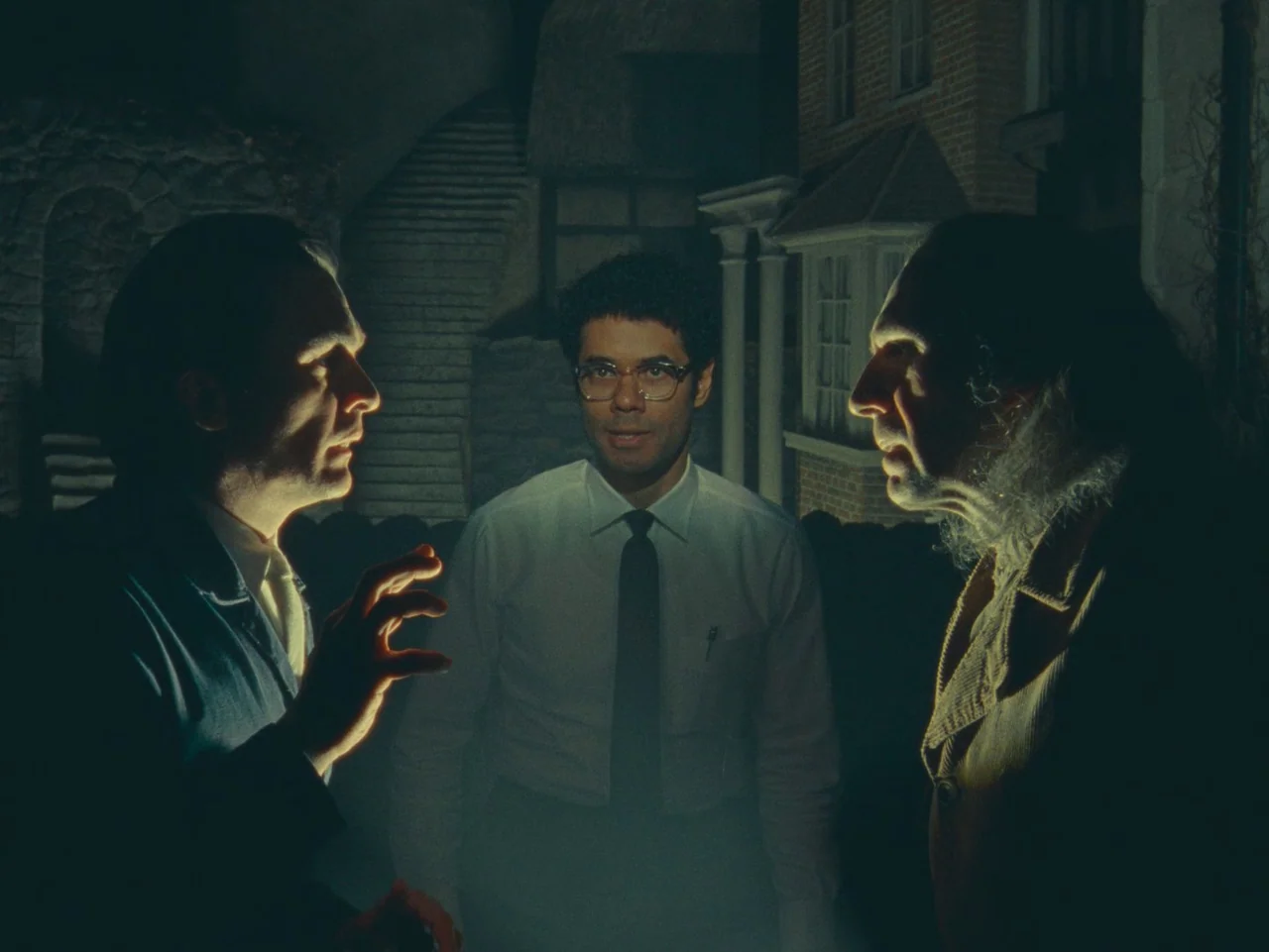 The Rat Catcher (2023) movie still: medium — Three men stand together in a dark space featuring miniature house facades behind them. The man on the…