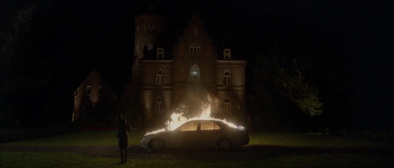 Nymphomaniac Vol 2 (2013) movie still: wide — A dark, expansive lawn sits before a large, multi-story brick manor house at night. A sedan is…