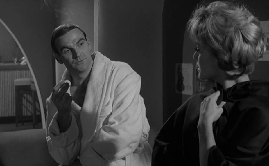 The Criminal (1960) movie still: medium — A dimly lit room with curved architectural elements is visible in the background. A man in a white robe…