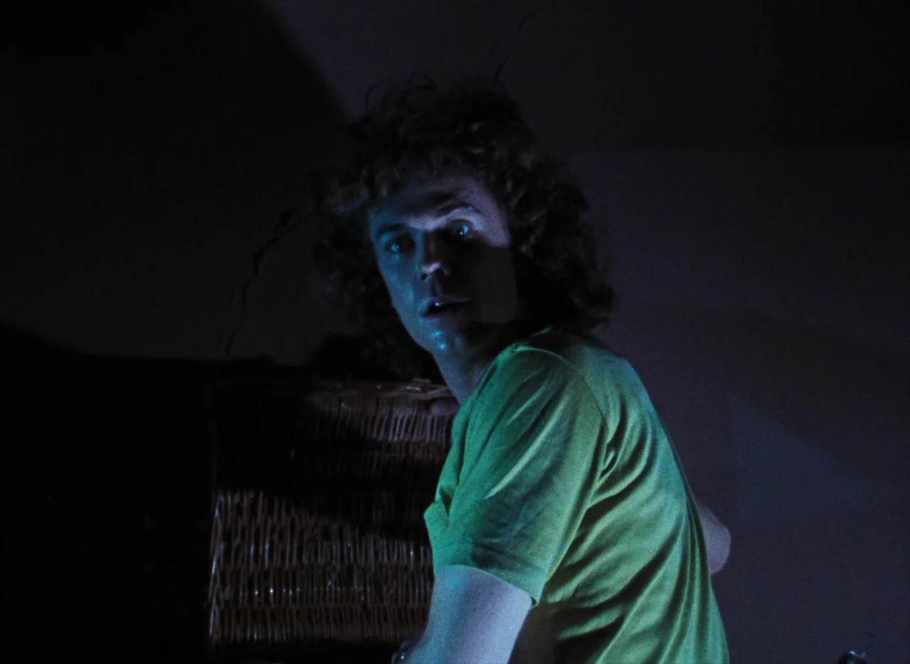 Basket Case (1982) movie still: medium — A dark, dimly lit indoor space with shadows occupying the majority of the frame. A young man with curly…