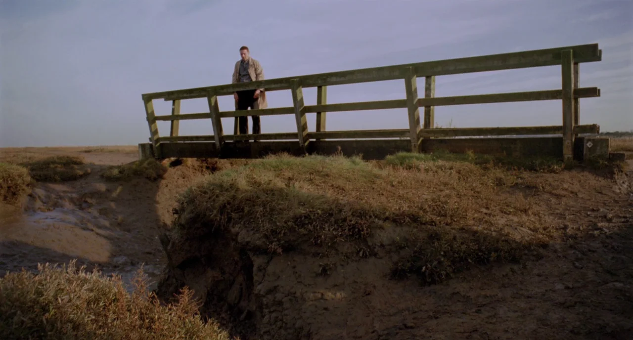 Possum (2018) movie still: wide — A rustic wooden footbridge spans a muddy tidal creek surrounded by low marshy vegetation. A man in a trench coat…