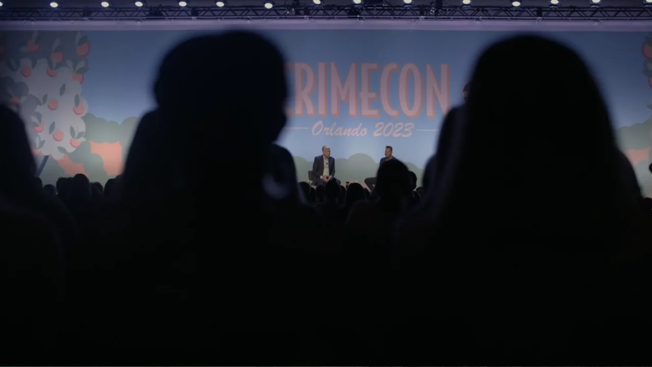 Predators (2010) movie still: wide — A large conference hall features a stage with a backdrop displaying branding for CrimeCon. Two men in dark…