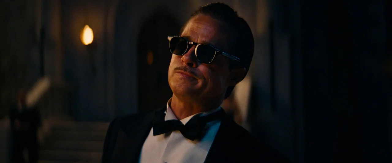 Babylon (2022) movie still: medium — A dimly lit ballroom with stone architectural features and soft warm wall lighting. A man wearing sunglasses…