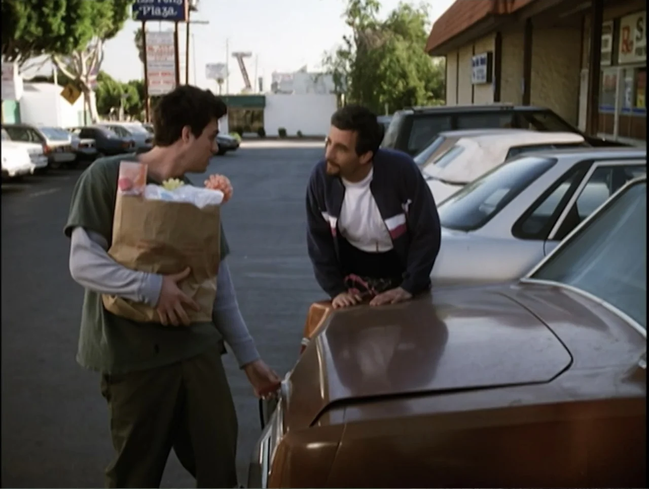 Apartment 12 (2001) movie still: medium — A sunlit parking lot in front of a strip mall with several parked cars. Two men stand beside a brown sedan…