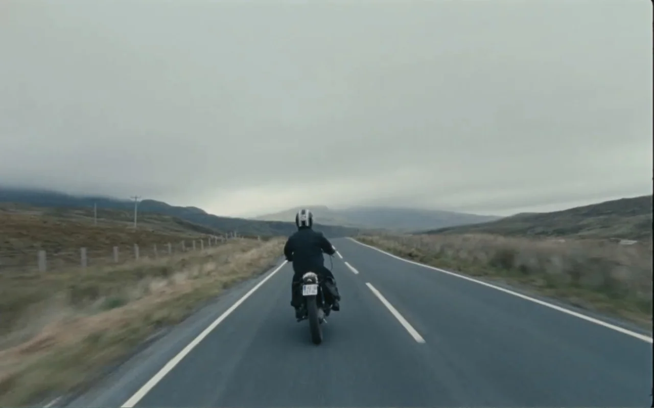 The Greasy Hands Preachers (2014) movie still: wide — A long paved road stretches through a hilly, desolate landscape under a gray sky. A lone…