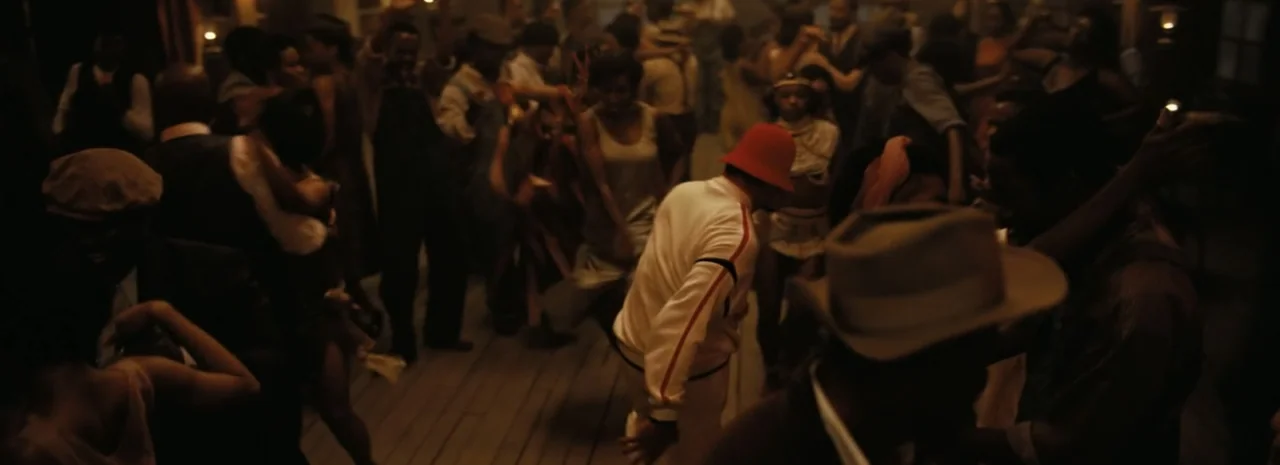 Sinners (2025) movie still: wide — A dimly lit, crowded dance hall features people dancing on a wooden floor at night. A man wearing a red hat and…