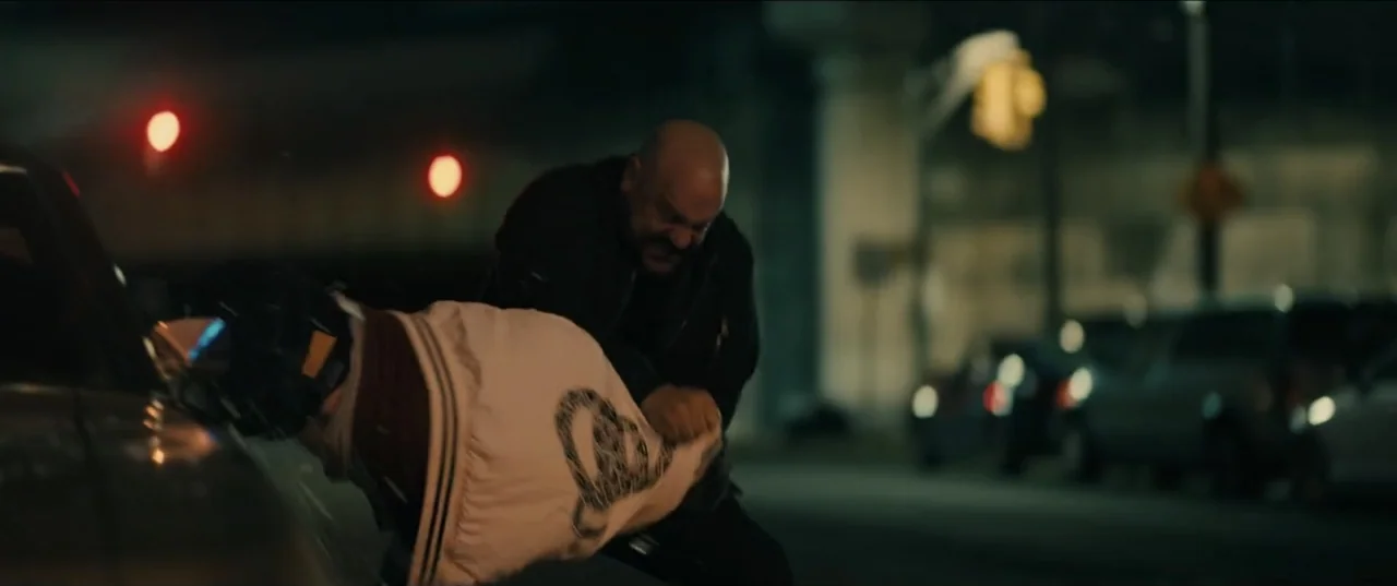 Guns Up (2025) movie still: medium — An urban street at night with parked cars and blurred background structures visible. A stocky bald man grips…