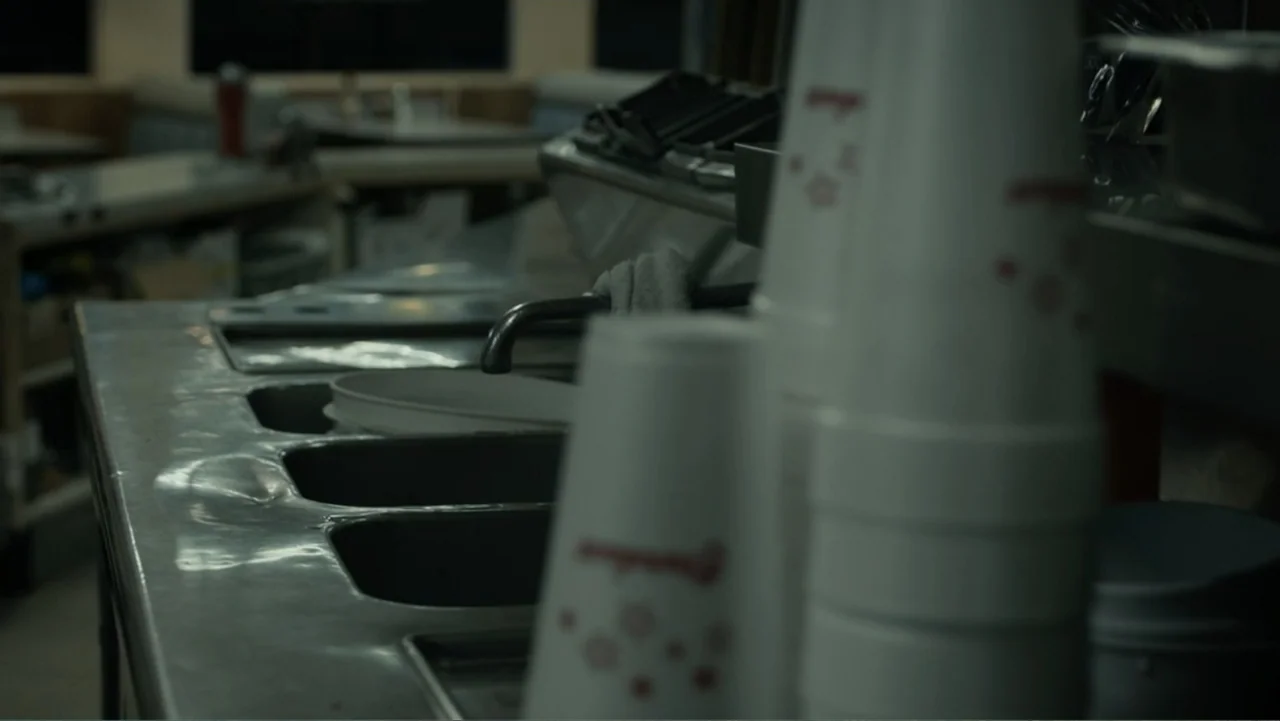 The Cleaner (2021) movie still: medium — A dark, commercial kitchen area features a weathered stainless steel triple-basin sink setup. Stacks of…