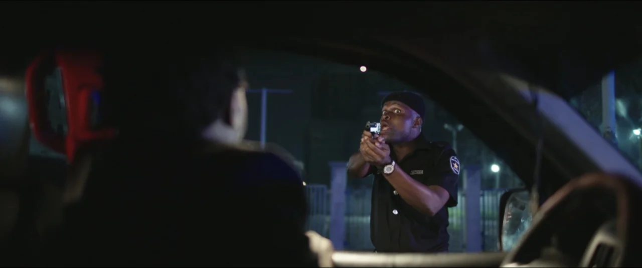 Saving Onome (2024) movie still: medium — An exterior night view from inside a car looking at a security barrier. A police officer points a handgun…
