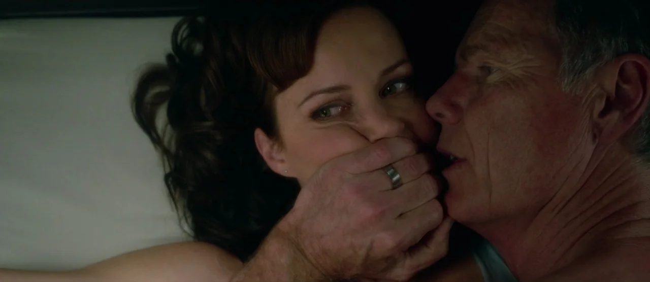 Gerald’s Game (2017) movie still: closeup — A dark, dimly lit bedroom setting focusing on two people in a bed. An older man presses his hand against…