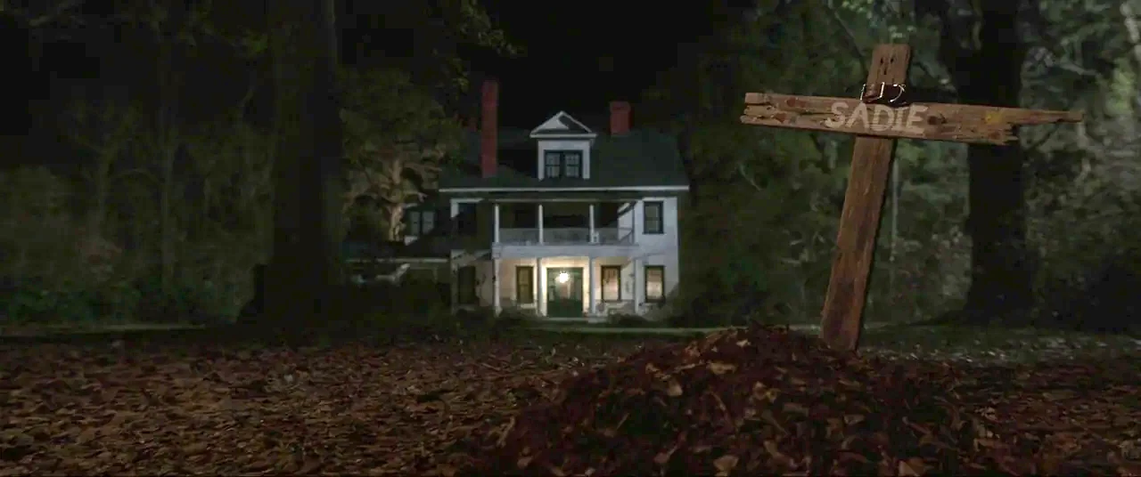 The Conjuring (2013) movie still: wide — A nighttime scene features a wooden cross marking a grave in a dark yard. A white two-story house with a…