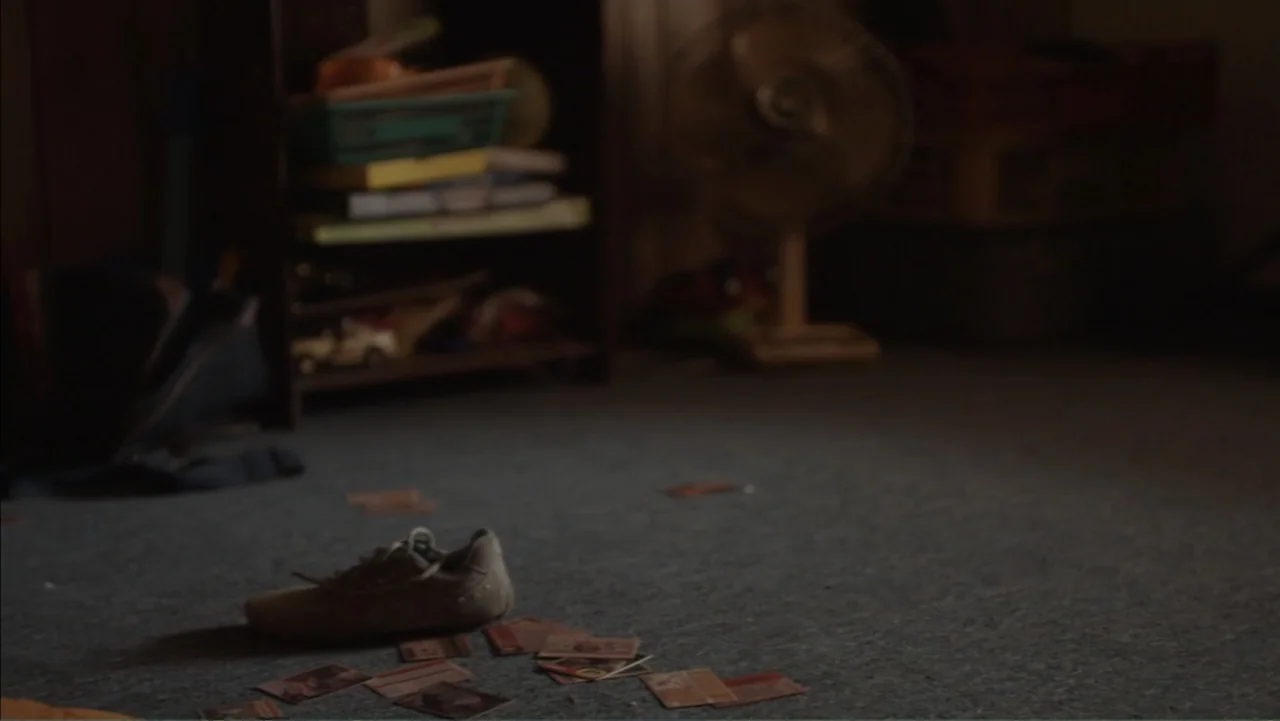Boy (2010) movie still: medium — A dimly lit room features a cluttered shelf and a standing fan behind items. A lone sneaker and several trading…