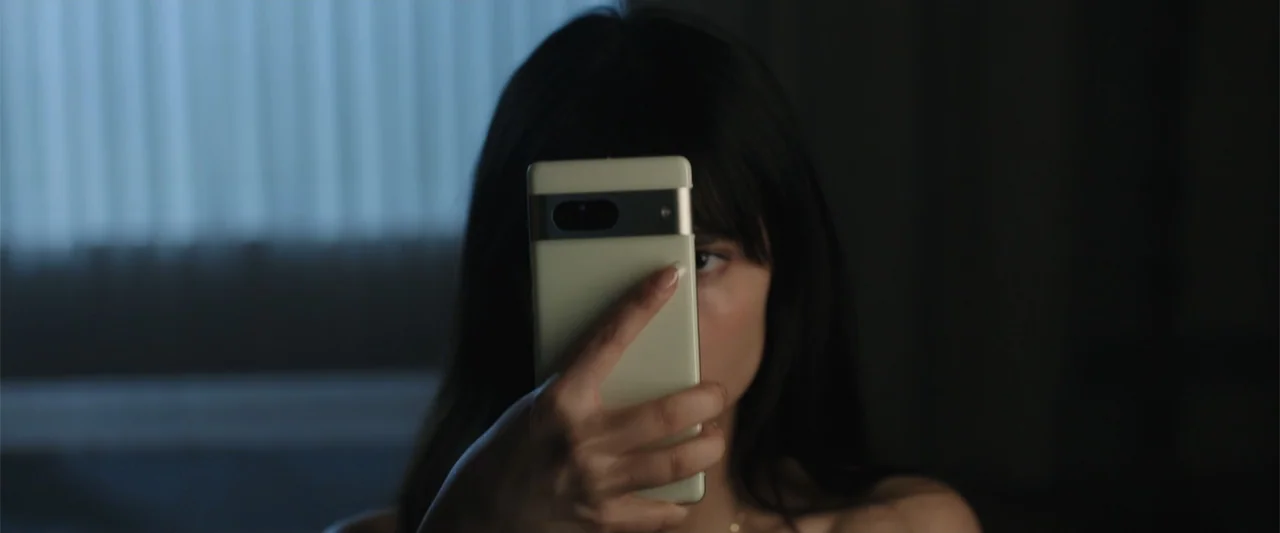 Trap House (2025) movie still: closeup — A dimly lit interior space contains a person partially obscured by a mobile device. The individual holds a…