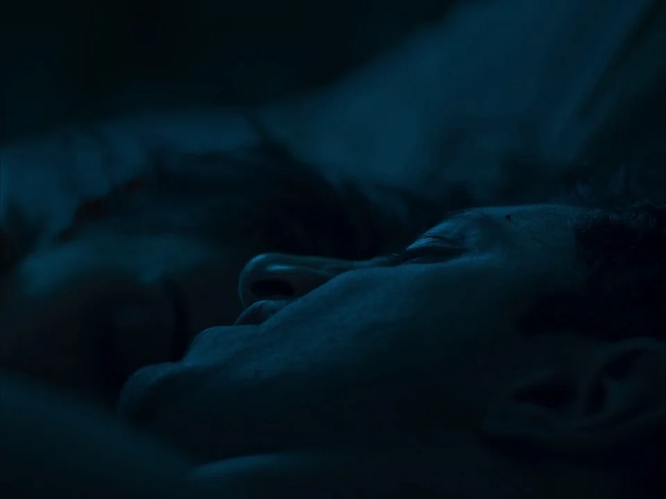 Mogul Mowgli (2020) movie still: closeup — A dark, dimly lit space showing two people resting close together. The focus is on the profile of one…