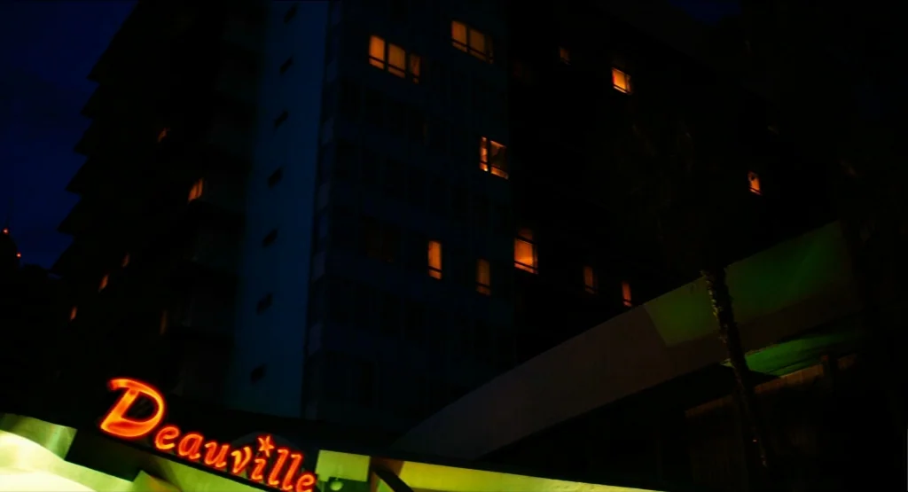Life During Wartime (2009) movie still: establishing — A dark hotel facade rises at night with scattered illuminated windows. An orange neon sign…