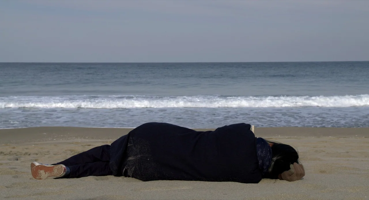 On The Beach At Night Alone (2017) movie still: fullbody — A wide beach features a shoreline with gentle waves meeting a sandy expanse. An elderly…