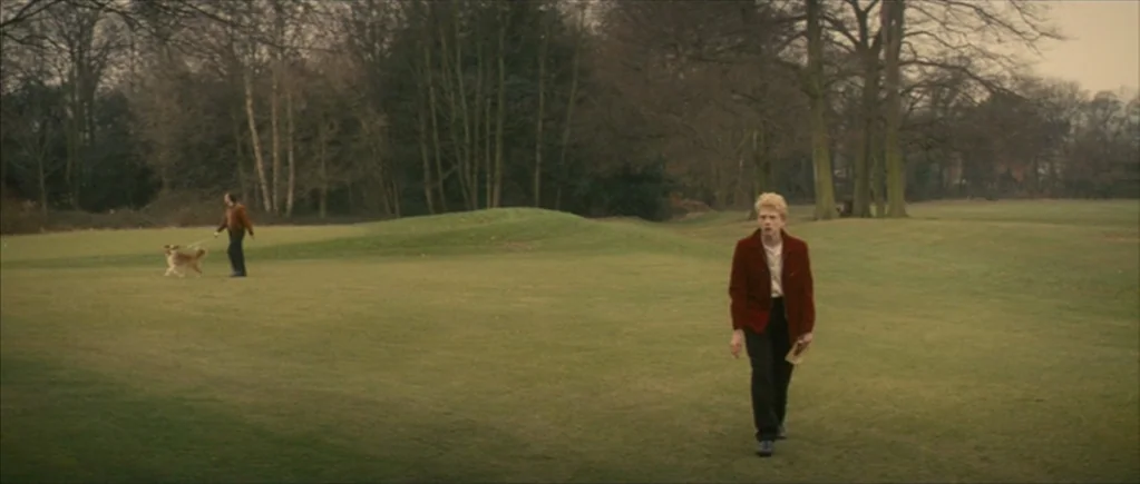 Nowhere Boy movie still: wide — A vast, open public field with short green grass and mature trees in background. A fair-skinned teenage boy in a…