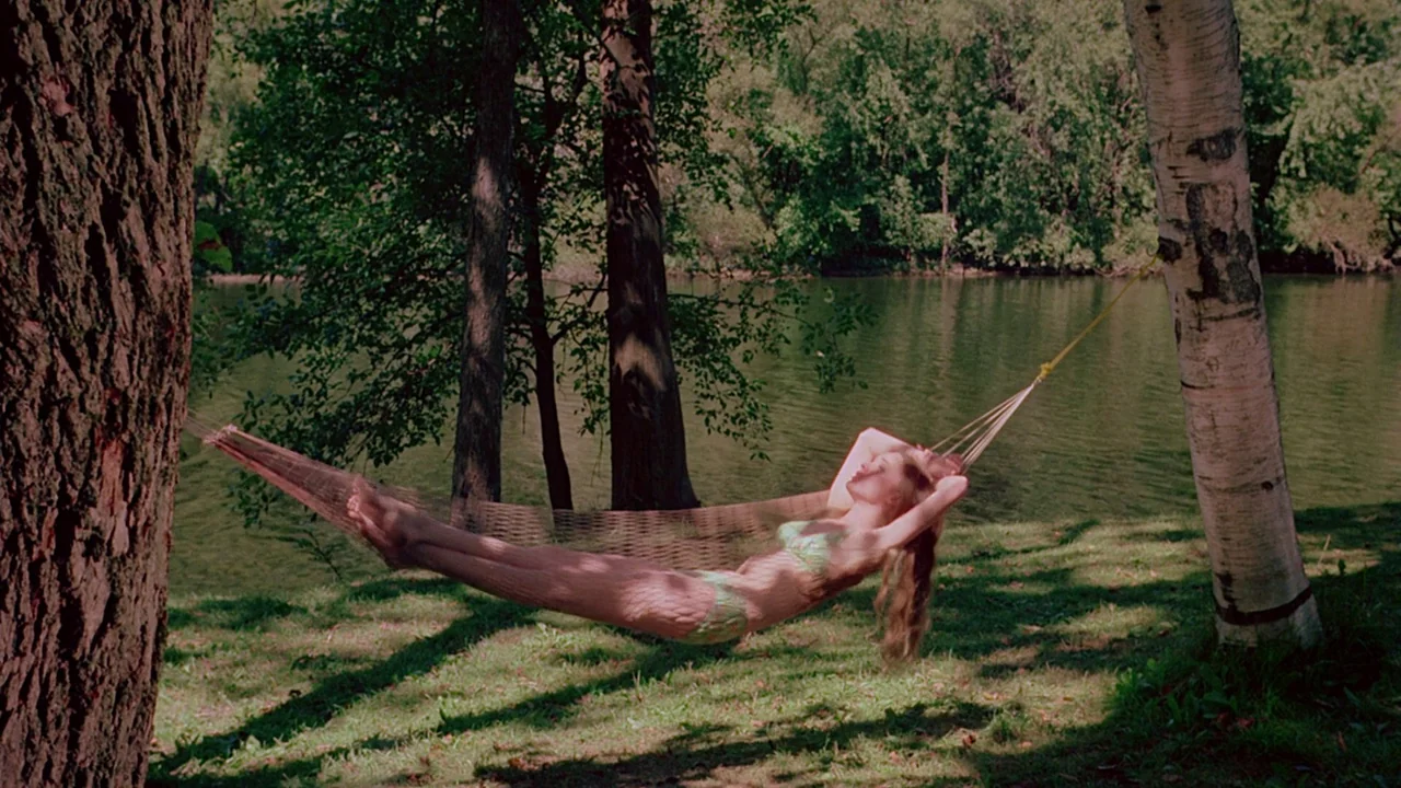 I Spit On Your Grave (1978) movie still: fullbody — A grassy riverbank shaded by mature trees features a mesh hammock strung between trunks. A woman…