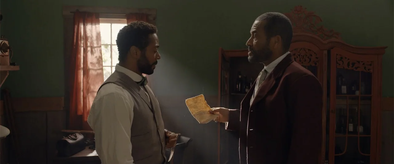 Ghosts of the Ozarks (2022) movie still: medium — A dimly lit 19th-century room features green walls and a large wooden cabinet. One man holds a…