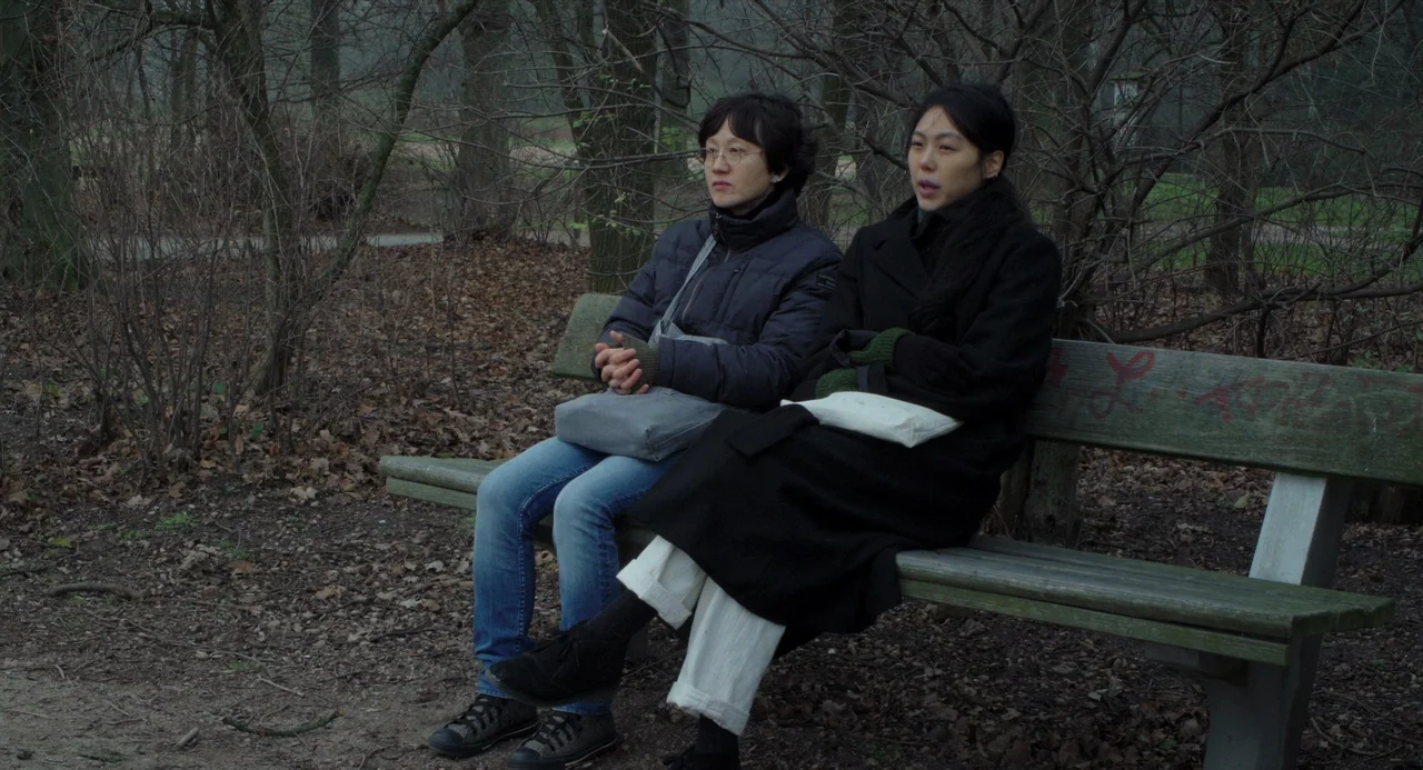 On The Beach At Night Alone (2017) movie still: medium — An outdoor park in late autumn with leafless trees and a dry, leaf-covered floor. Two…