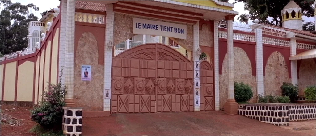 White Material (2009) movie still: wide — An ornate entrance gate features a sign reading LE MAIRE TIENT BON. Political posters are plastered onto…