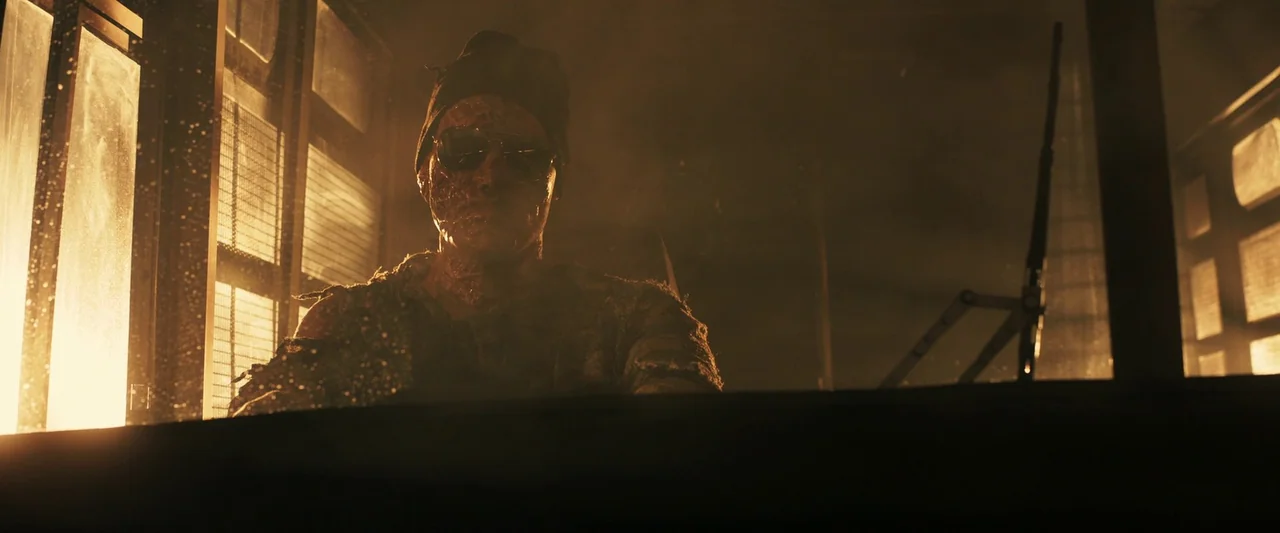 Prisoners of the Ghostland (2021) movie still: medium — A dimly lit guard booth filled with smoke and amber-toned light at night. A scarred man…