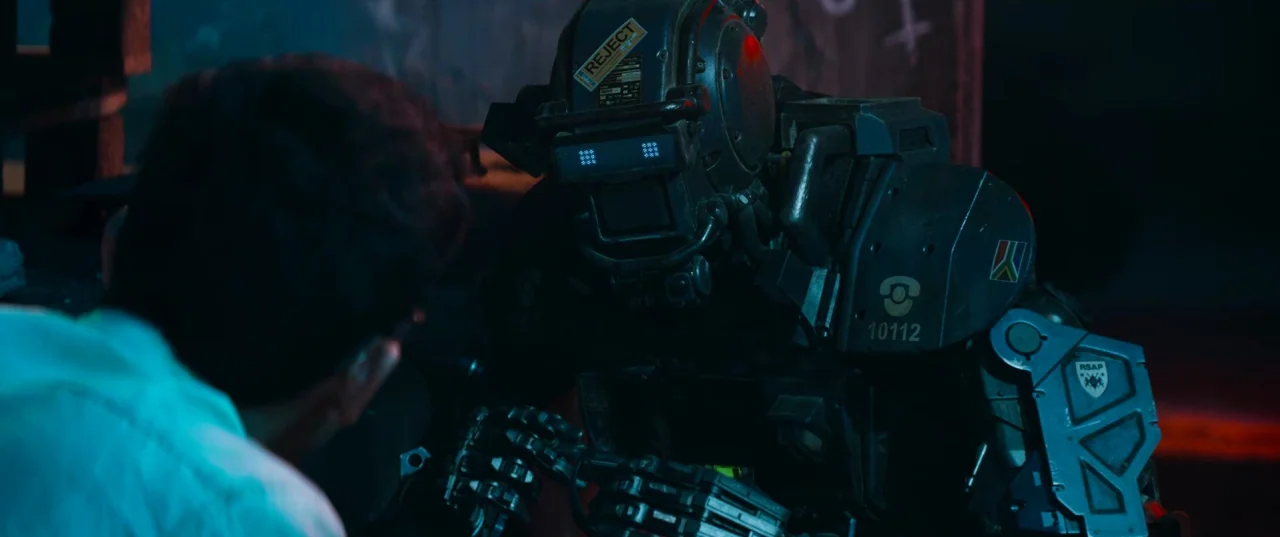 Chappie (2015) movie still: medium — A dimly lit industrial workspace contains a mechanical robot with prominent blue optical sensors. The robot…
