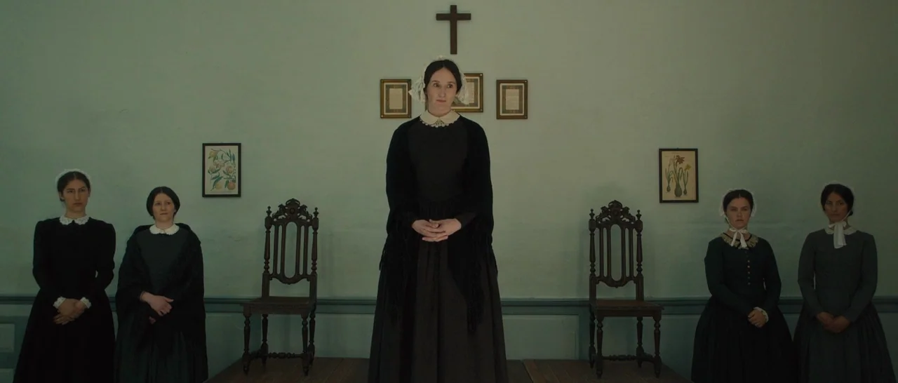 A Quiet Passion (2016) movie still: fullbody — Five women stand in a symmetrical line against a pale green wall. A small wooden cross is mounted…