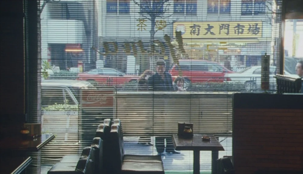 Gozu (2003) movie still: wide — An empty diner booth sits behind a window covered by thin horizontal blinds. Outside, the blurred street features a…