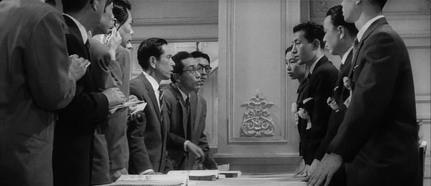 The Bad Sleep Well (1960) movie still: wide — A formal interior space features a table separating two groups of men in suits. The men lean across…