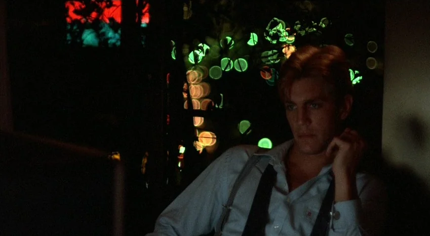 The Coca-Cola Kid (1985) movie still: medium — A dimly lit room with an out-of-focus background showing vibrant green and orange bokeh patterns. A…