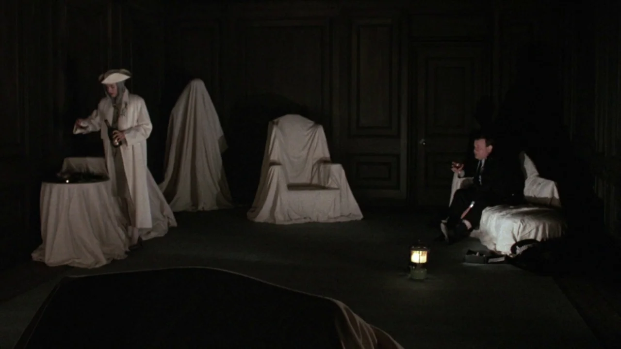 The Lair of the White Worm (1988) movie still: wide — A dark, sparsely furnished room features wood paneling and furniture covered in white cloth. A…