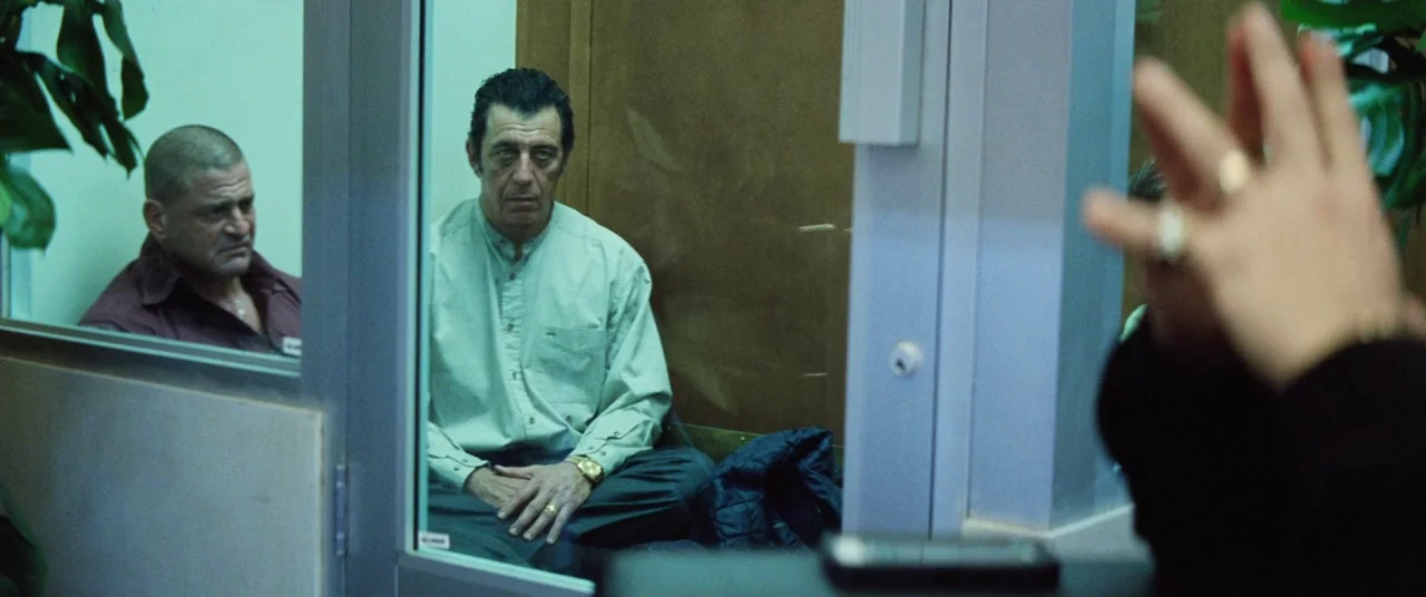 Uncut Gems (2019) movie still: medium — Two men sit within a small glass-walled room in a sterile building interior. They appear to be waiting while…