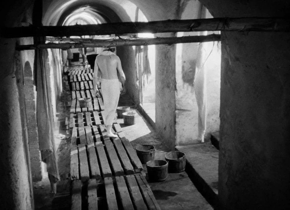 Othello (1952) movie still: fullbody — An arched stone corridor contains a raised wooden walkway and several discarded buckets. A shirtless man…