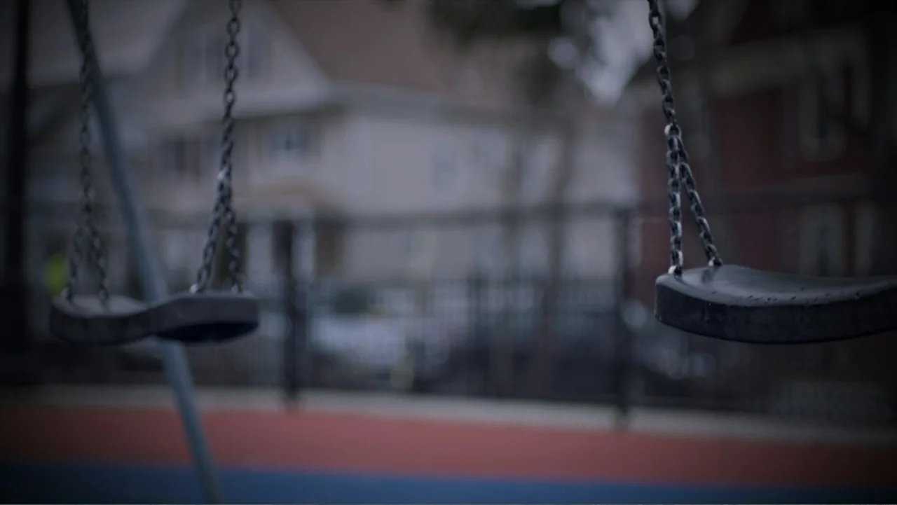 What Happened at Fells Acres? (2023) movie still: medium — Two black rubber swings hang in the foreground of a blurred playground. A chain-link…