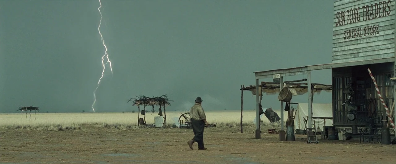 The Proposition (2005) movie still: wide — A rural settlement features a wooden general store beside a vast expanse of dry grass. A single man walks…