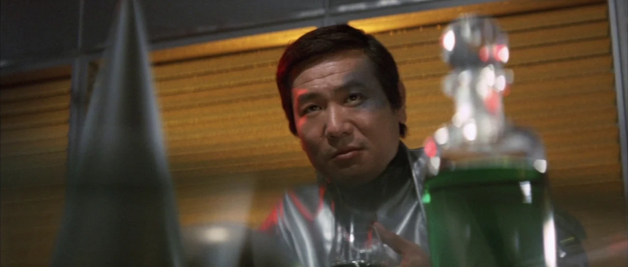 Godzilla Vs Mechagodzilla (1974) movie still: medium — A dimly lit room features horizontal blinds in the background and metallic wall panels. A man…
