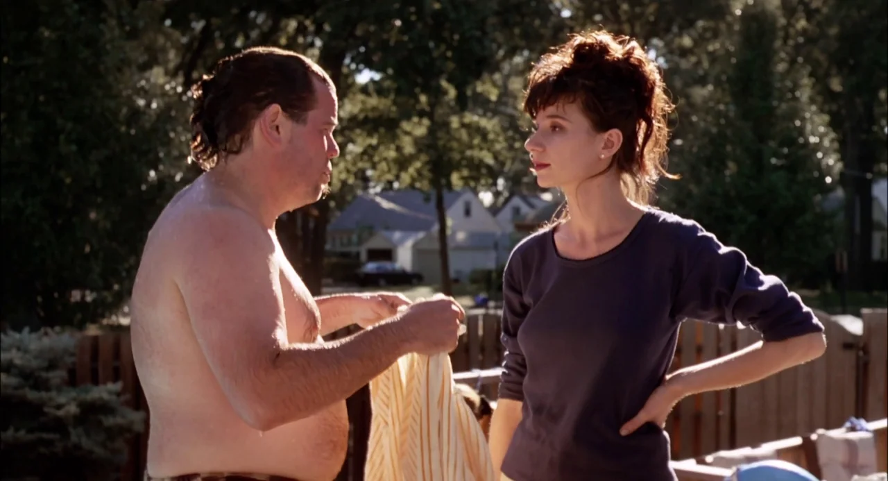 Trees Lounge (1996) movie still: medium — A suburban backyard with a wooden fence and leafy trees in the background. A shirtless man holding a…