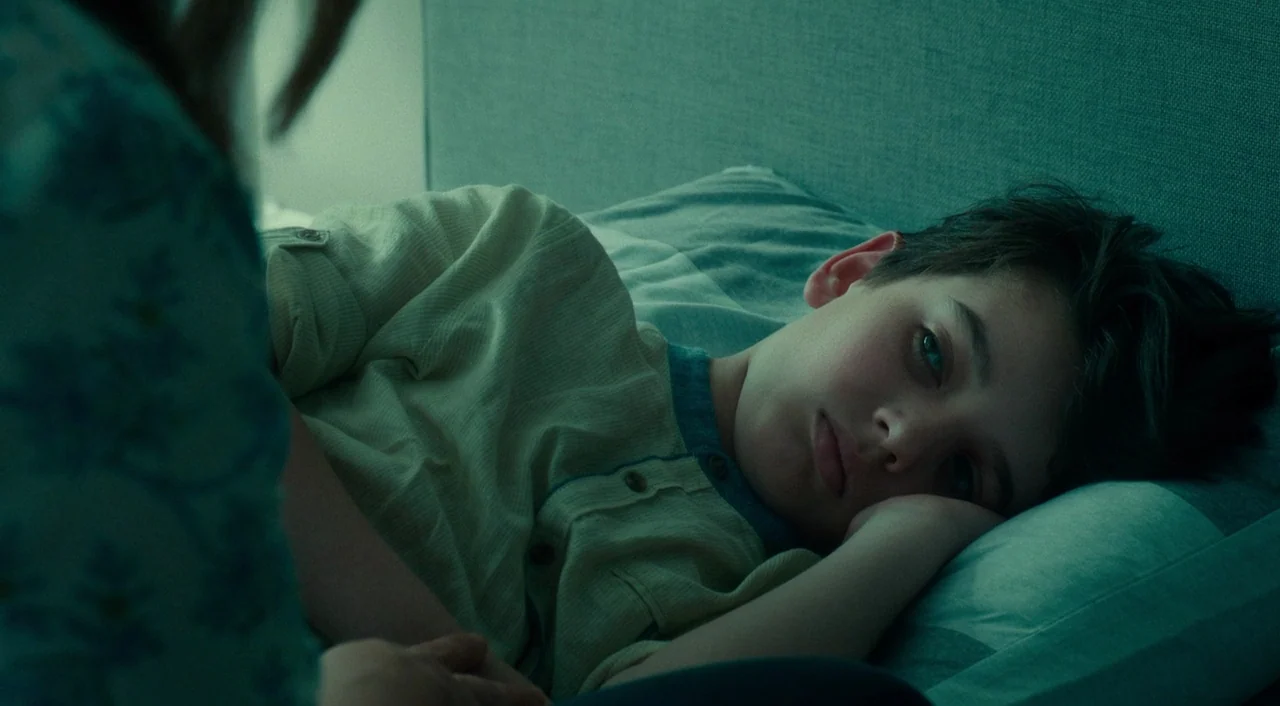 Wiener-Dog (2016) movie still: medium — A young boy rests his head on a pillow in a dimly lit room. He is positioned in the bed while another person…