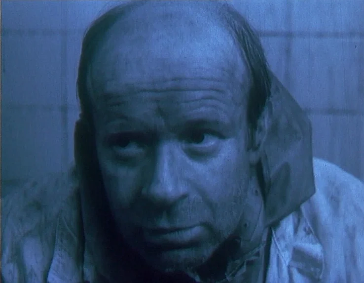 Dead Man’s Letters (1986) movie still: closeup — A close-up against a wall of pale square tiles in a dimly lit space. A middle-aged man with…