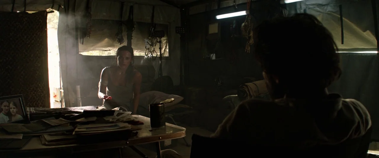 Tomb Raider (2018) movie still: medium — A cluttered tent interior has gear hanging from the walls and a central table. A woman sits facing the…