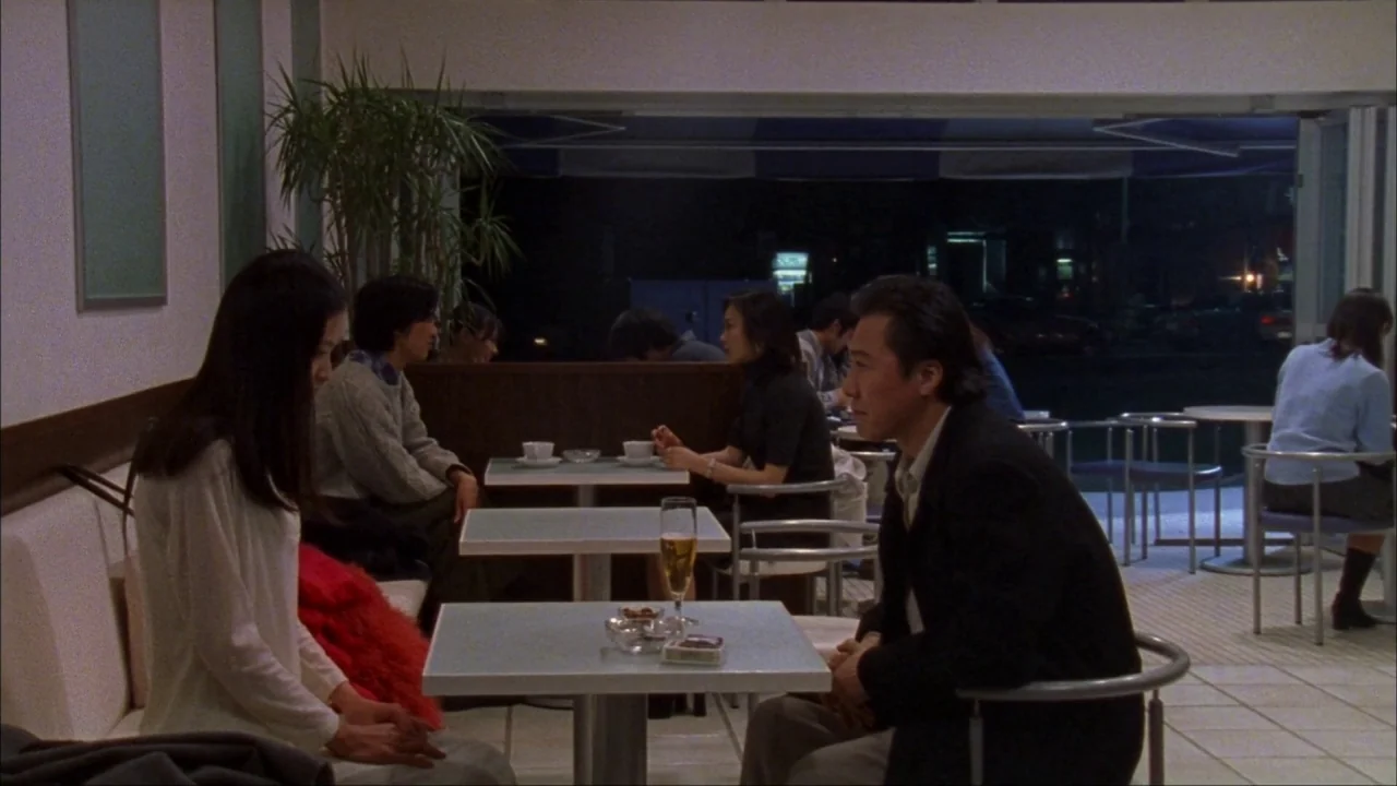Audition (1999) movie still: medium — A quiet cafe interior features tiled floors and several tables at night. A man and a woman sit facing each…