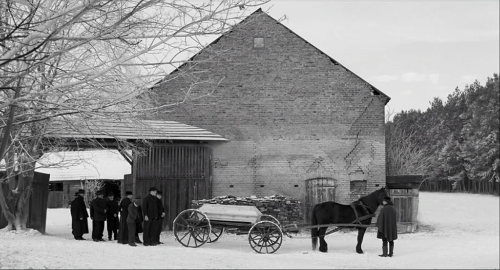 The White Ribbon (2009) movie still: wide — A group of men in dark coats stand in a snow-covered farmyard. A horse pulls a cart with a coffin in…